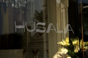 Husala logo etched on glass reflecting an interior with a chandelier and greenery in soft focus