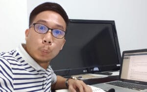 Person in striped polo shirt and glasses making a playful face while sitting at a desk with monitor and laptop